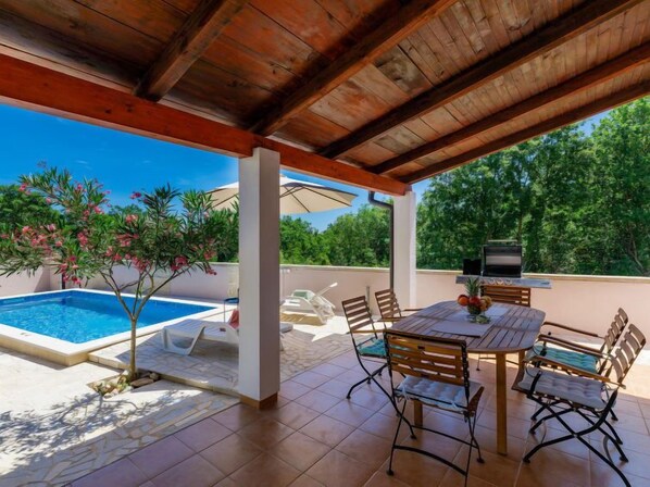 Un espacio al aire libre techado con piscina, una mesa con sillas y una parrilla.