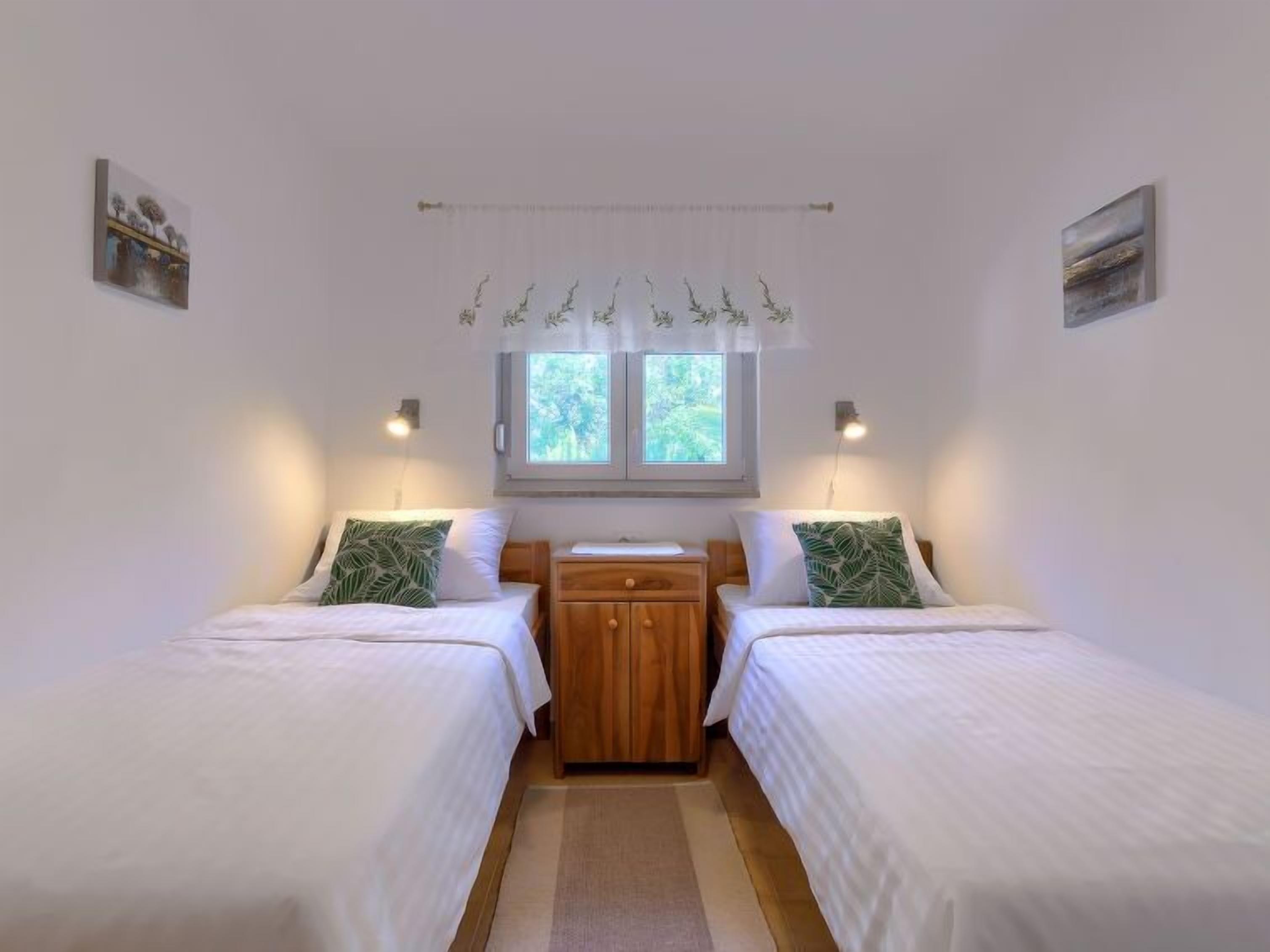 A room with two single beds, a wooden nightstand, a window with curtains, and wall-mounted lights.