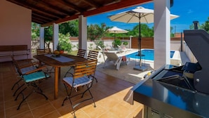 An outdoor patio with a pool, wooden furniture, and a grill. - Maslina by Interhome (Pula/Duga uvala)