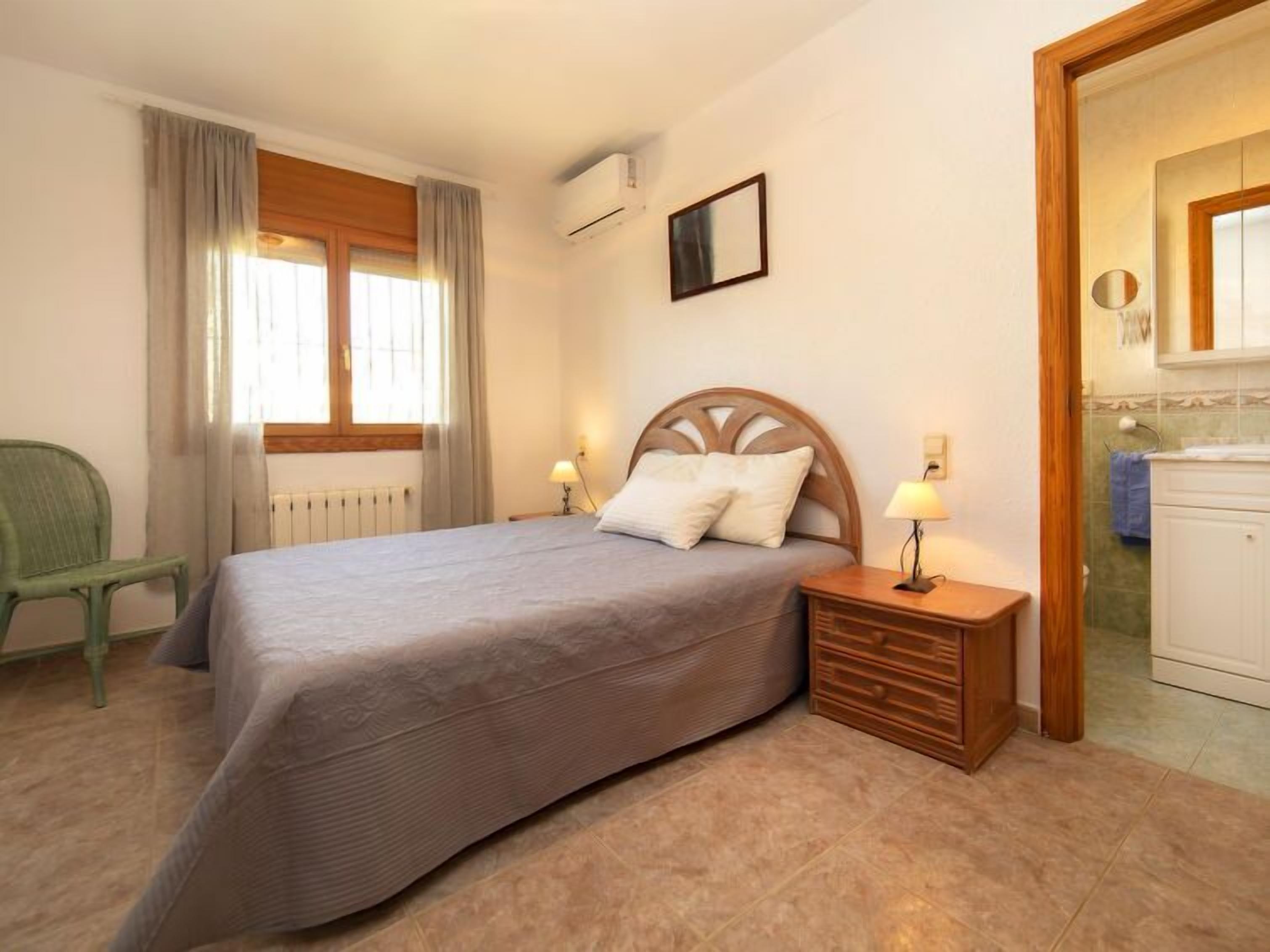 A bedroom with a bed, two bedside tables with lamps, a chair, a window with curtains, an air conditioning unit, and a bathroom visible through an open door.
