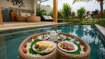 A poolside breakfast setup with a wicker table, plates of food, a teapot, and a glass of drink.