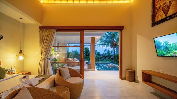 A modern hotel room with a large bed, wicker furniture, a TV, and a view of a pool and palm trees. - Arta Private Villa (Ubud)