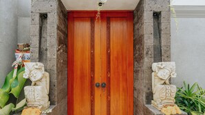 A wooden door with two stone statues on either side, a small shelf with items, and a stone wall. - Arta Private Villa (Ubud)