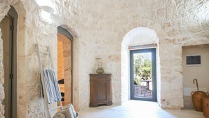 A rustic stone interior with a wooden cabinet, a metal bucket with towels, and a view through an open door. - TD Trulli Albapetra Pool Olive Trees (Polignano a Mare)