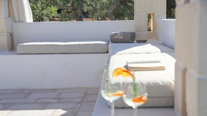 A sunlit outdoor seating area with a cushioned bench and a small table, a rolled towel, and two glasses of a clear beverage. - TD Trulli Albapetra Pool Olive Trees (Polignano a Mare)