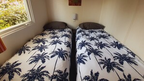 Ein kleines Zimmer mit zwei Einzelbetten, einem Fenster mit Blick auf Grünflächen, einem an der Wand befestigten Regal und einer Wandleuchte.
