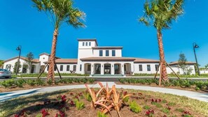 A two-story building with a central entrance, flanked by palm trees, and a landscaped garden with red flowers in the foreground. - Stunning Lake View! Themed Bedrooms With Comfortable and Sophisticated Decor (Kissimmee)