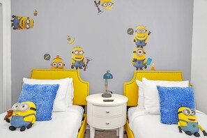 A child's bedroom with two beds, each with a yellow headboard and white bedding. There are two blue textured pillows, a white bedside table with a lamp, and a small drawer. Minion-themed decorations are visible on the walls and furniture. - Stunning Lake View! Themed Bedrooms With Comfortable and Sophisticated Decor (Kissimmee)
