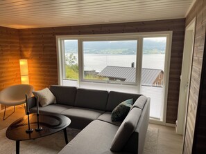 Ein modernes Wohnzimmer mit einer Eckcouch, einem runden Couchtisch und einer Stehlampe. Große Fenster bieten Blick auf einen See und Gebäude.