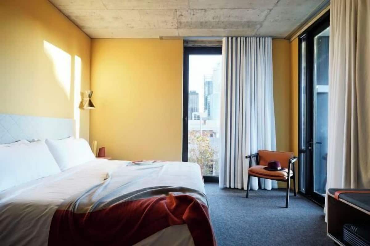 Habitación de hotel con cama, una silla, ventana con cortinas y vistas a edificios.