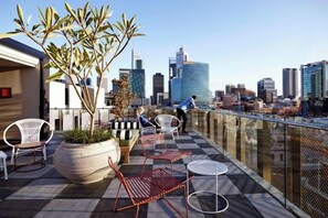 A rooftop terrace with seating, a large potted plant, and a city skyline view. - Alex Hotel (Northbridge)