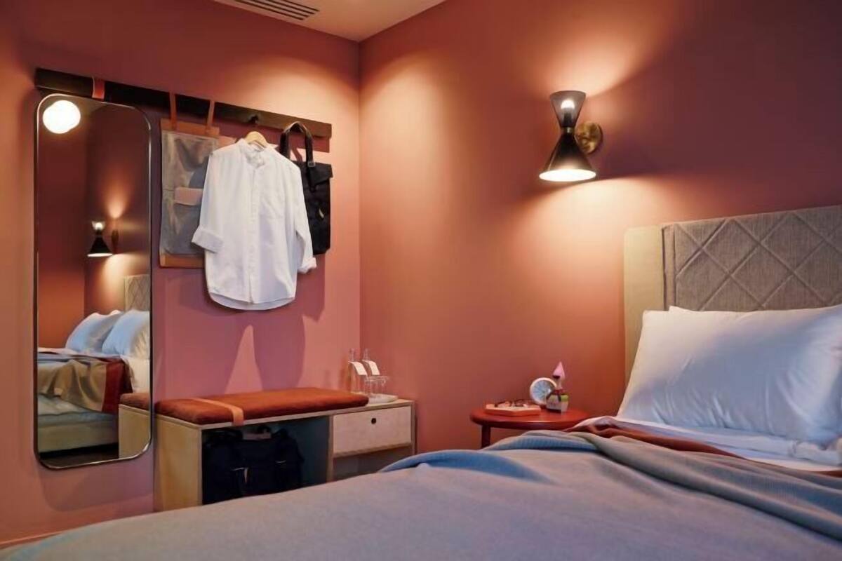 Habitación de hotel con cama, mesita de noche con lámpara, espejo y perchero de pared con una camisa blanca y un bolso negro.