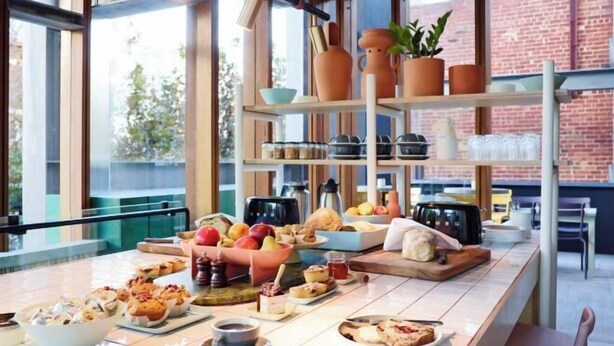 A breakfast buffet with various food items and beverages, a coffee station, and a view of a brick building through large windows. - Alex Hotel (Northbridge)