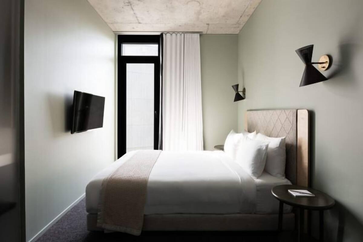 Habitación de hotel moderna con una cama grande, un televisor de pantalla plana montado en la pared, una mesita de noche y una ventana con cortinas.