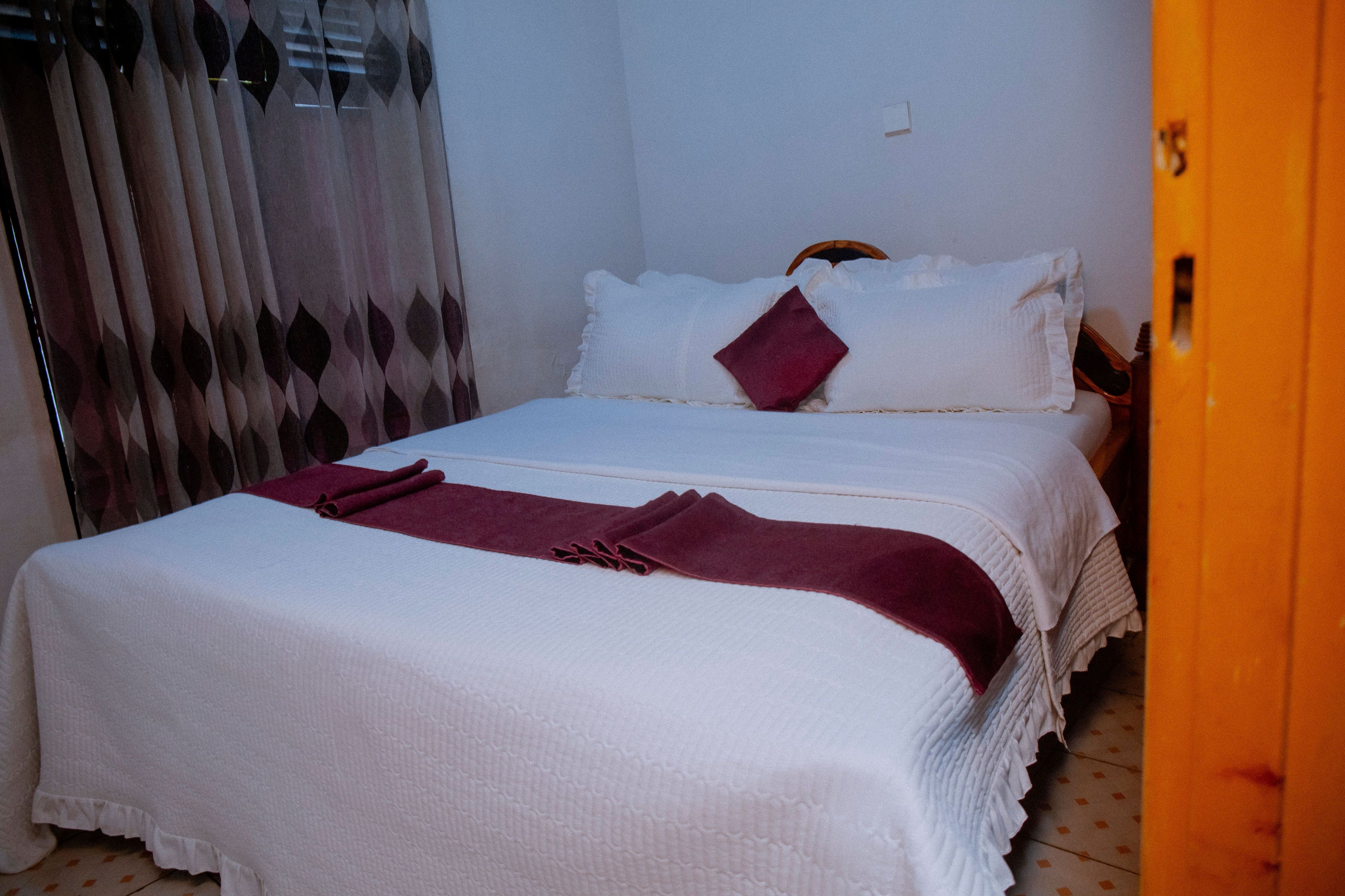 A neatly made bed with white bedding and a red throw blanket, a wooden headboard, and a curtain with a geometric pattern.