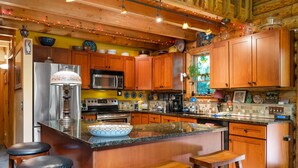 A kitchen with wooden cabinets, a stainless steel refrigerator, a microwave, and a large island with stools. - Beautiful Log Cabin | Wooded | Lake Access | Interlochen Arts Academy (Interlochen)