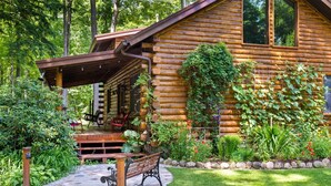 A log cabin with a covered porch, outdoor seating, and a garden with various plants and flowers. - Beautiful Log Cabin | Wooded | Lake Access | Interlochen Arts Academy (Interlochen)