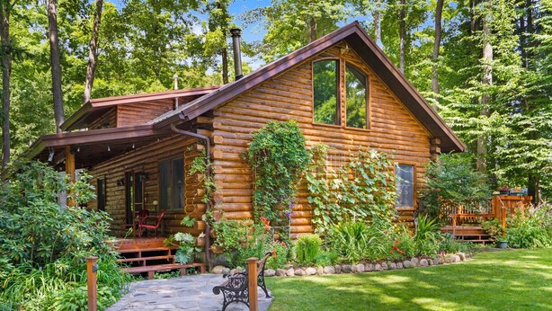 A log cabin with a porch, surrounded by lush greenery and a stone pathway. - Beautiful Log Cabin | Wooded | Lake Access | Interlochen Arts Academy (Interlochen)