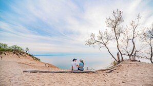 A couple sitting on a log on a sandy beach with a calm body of water and sparse vegetation in the background. - Beautiful Log Cabin | Wooded | Lake Access | Interlochen Arts Academy (Interlochen)