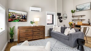 Un salon moderne avec un canapé gris, une commode en bois et une table basse noire. Un grand téléviseur est fixé au mur et affiche une photo de groupe.