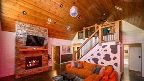 A cozy living room with a stone fireplace, a large orange sofa, and a wooden staircase. - 2-Cabin Retreat| Pool Table | Hot Tubs | Mini Golf (Broken Bow)