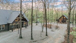 A rustic cabin with a sloped roof, surrounded by tall trees and a gravel driveway. - 2-Cabin Retreat| Pool Table | Hot Tubs | Mini Golf (Broken Bow)