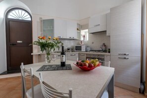 A modern kitchen with white cabinets, a dining table with chairs, a vase with flowers, and a bowl of fruit.