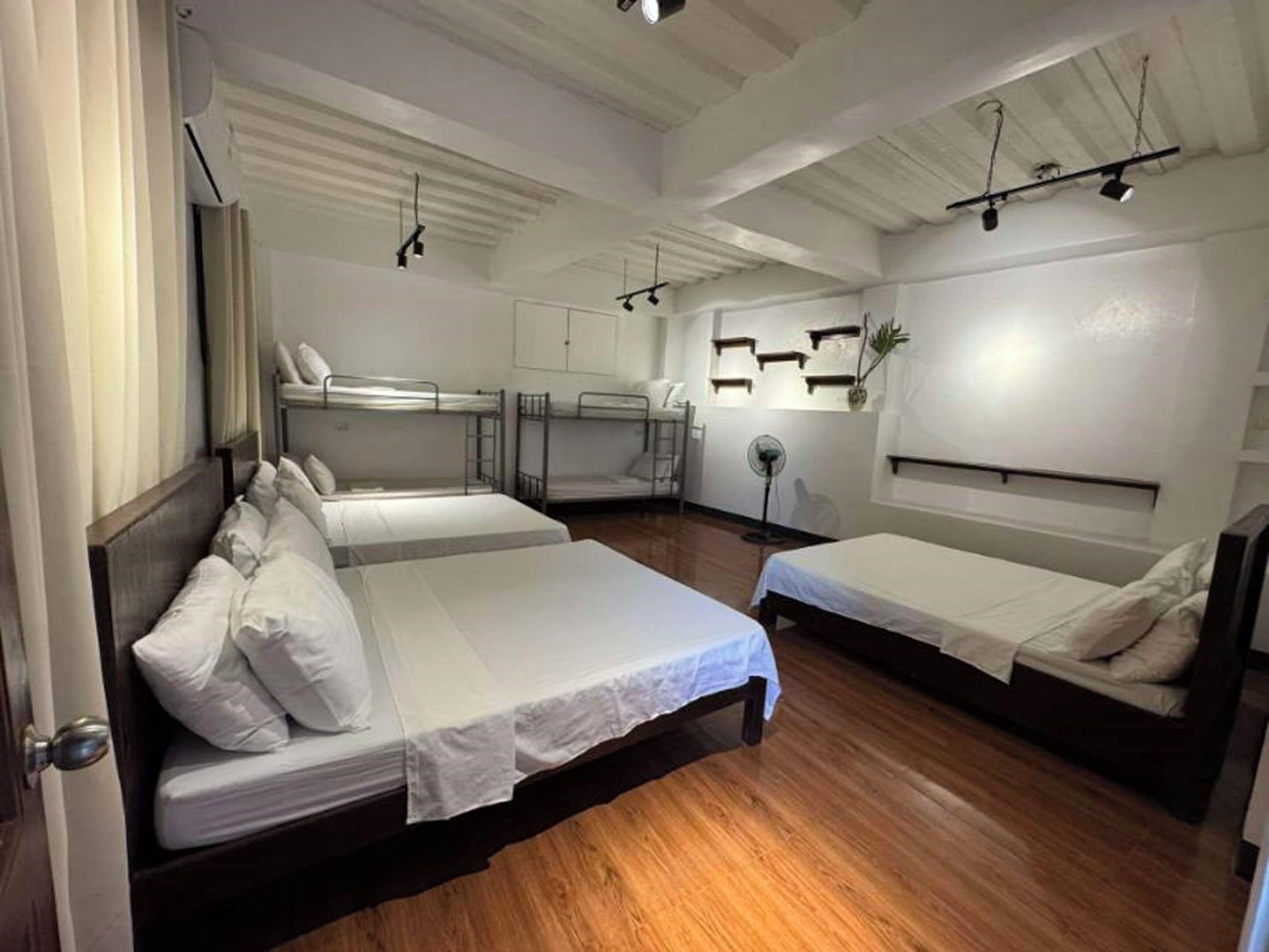 Habitación de hotel con dos camas individuales, una litera y suelo de madera, además de un ventilador de techo.
