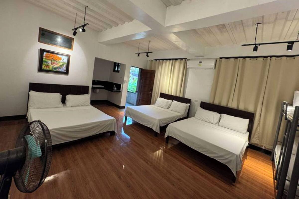 Een hotelkamer met drie bedden, een ventilator, een schilderij aan de muur en een raam met gordijnen.