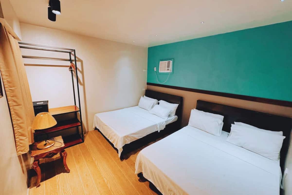 Een hotelkamer met twee enkele bedden, een houten vloer, een wandairconditioner en een tafeltje met een lamp.