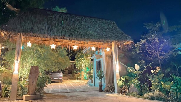 A thatched-roof structure with star-shaped lights, surrounded by lush greenery and a paved entrance.