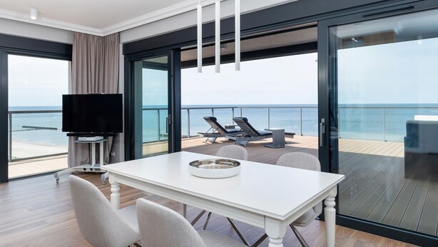 Ein modernes Zimmer mit einem weißen Tisch, Stühlen, einem Fernseher und einem Balkon mit Meerblick.