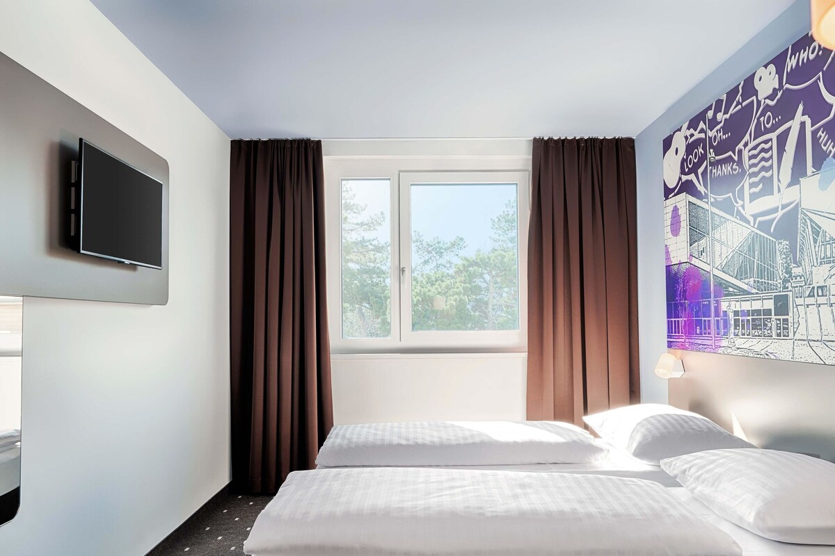 Une chambre d’hôtel avec deux lits, un téléviseur à écran plat fixé au mur, une fenêtre avec des rideaux et une fresque représentant un paysage urbain sur le mur.