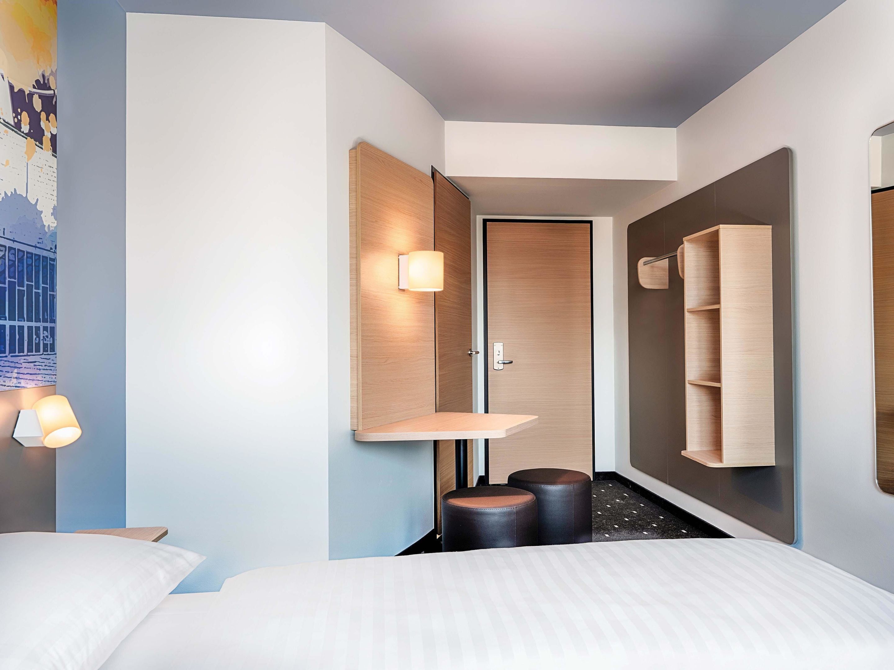 A modern hotel room with a bed, bedside tables, a wall-mounted shelf, a door, and a small stool.