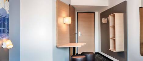 A modern hotel room with a bed, bedside tables, a wall-mounted shelf, a door, and a small stool.