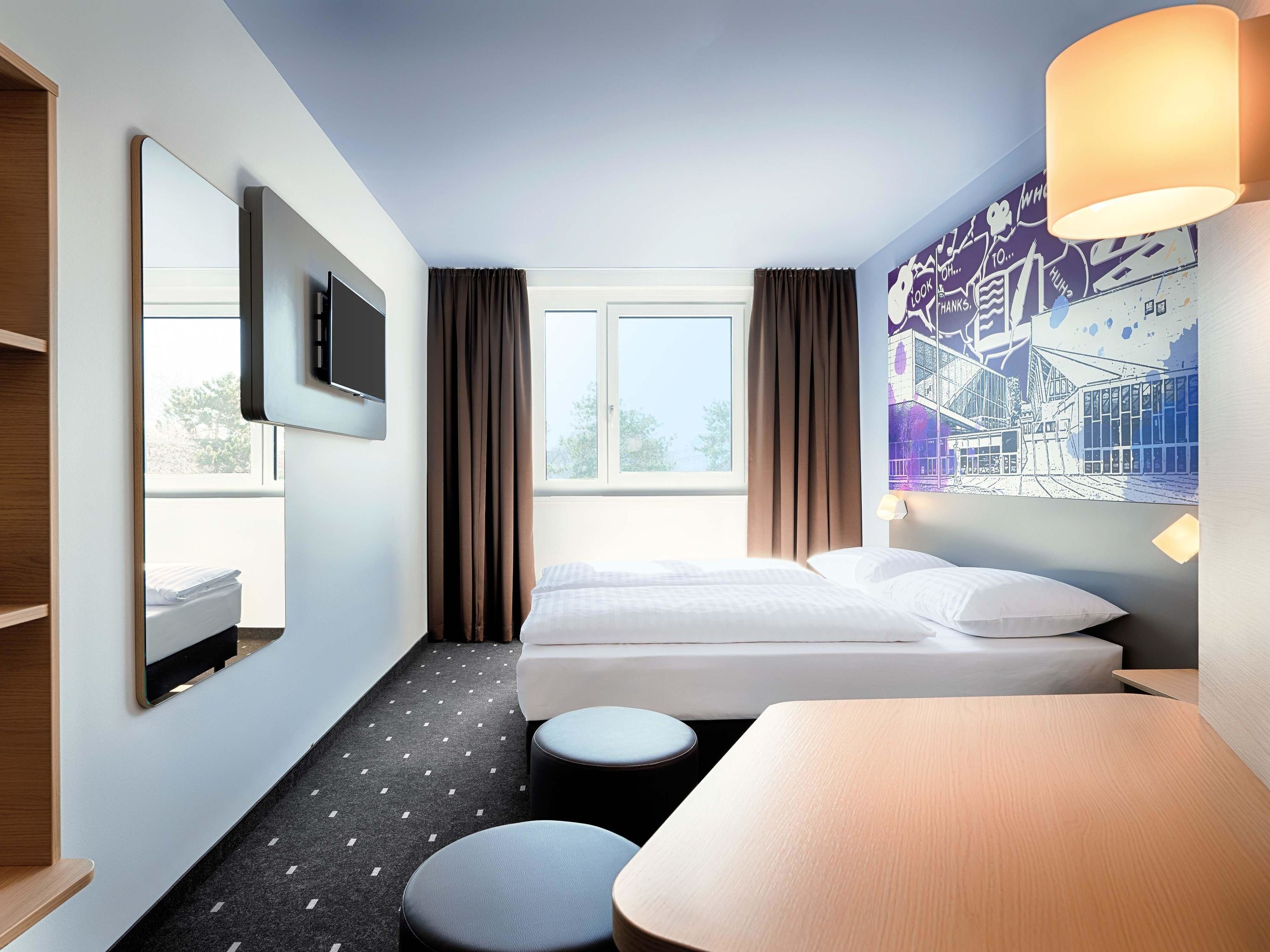 A hotel room with a bed, a TV, a desk, and a window with curtains.