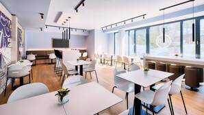Un espace repas moderne avec des tables et des chaises blanches, une cuisine en arrière-plan et de grandes fenêtres qui laissent entrer la lumière naturelle.