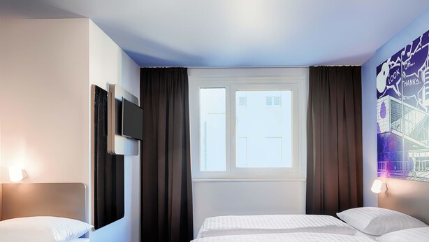 Une chambre d’hôtel avec deux lits, une télévision, une fenêtre avec des rideaux et un mur orné d’une fresque représentant un paysage urbain.
