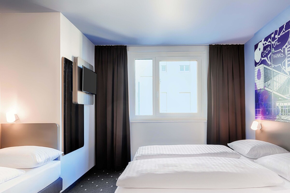 Une chambre d’hôtel avec deux lits, une télévision, une fenêtre avec des rideaux et un mur orné d’une fresque représentant un paysage urbain.