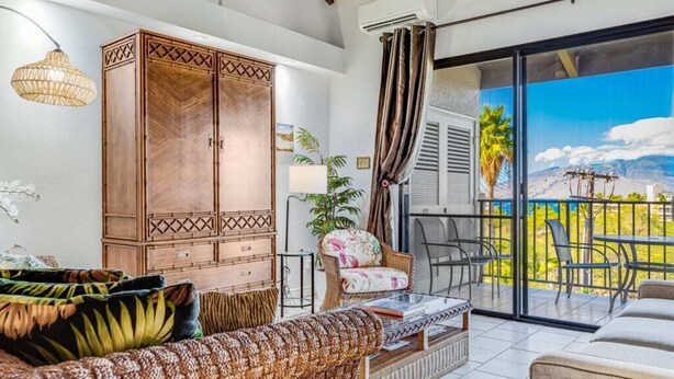 A spacious living area with a large wooden cabinet, a wicker sofa, and a balcony with a view of palm trees and mountains. - Roomy 2-Story w/Updated Kitchen–Kamaole Sands 4403 (KIHEI)