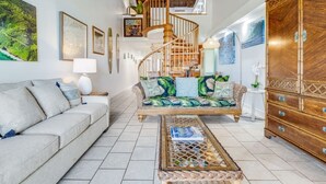 A spacious living room with a high ceiling, a wooden staircase, a sofa with tropical cushions, a coffee table, and a large wooden cabinet. - Roomy 2-Story w/Updated Kitchen–Kamaole Sands 4403 (KIHEI)