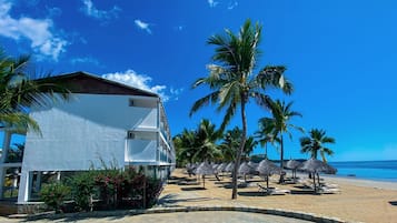 Un edificio blanco con balcón, palmeras y sombrillas de playa.