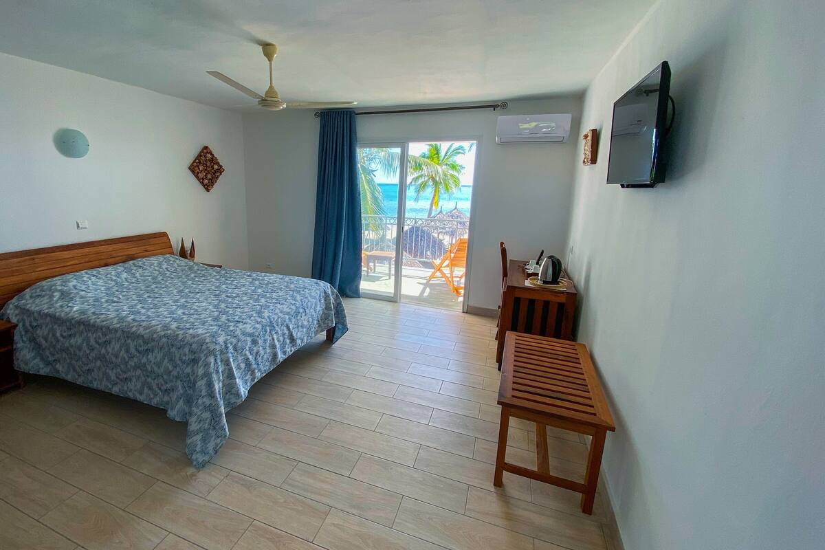 Ein Schlafzimmer mit einem Bett, einem Nachttisch aus Holz, einem an der Wand befestigten Fernseher und einem Balkon mit Blick auf Palmen.