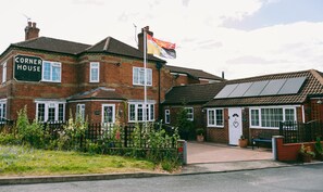 A corner house with a sign reading "Corner House," a flagpole with a flag, and a small garden with flowers and a fence.
