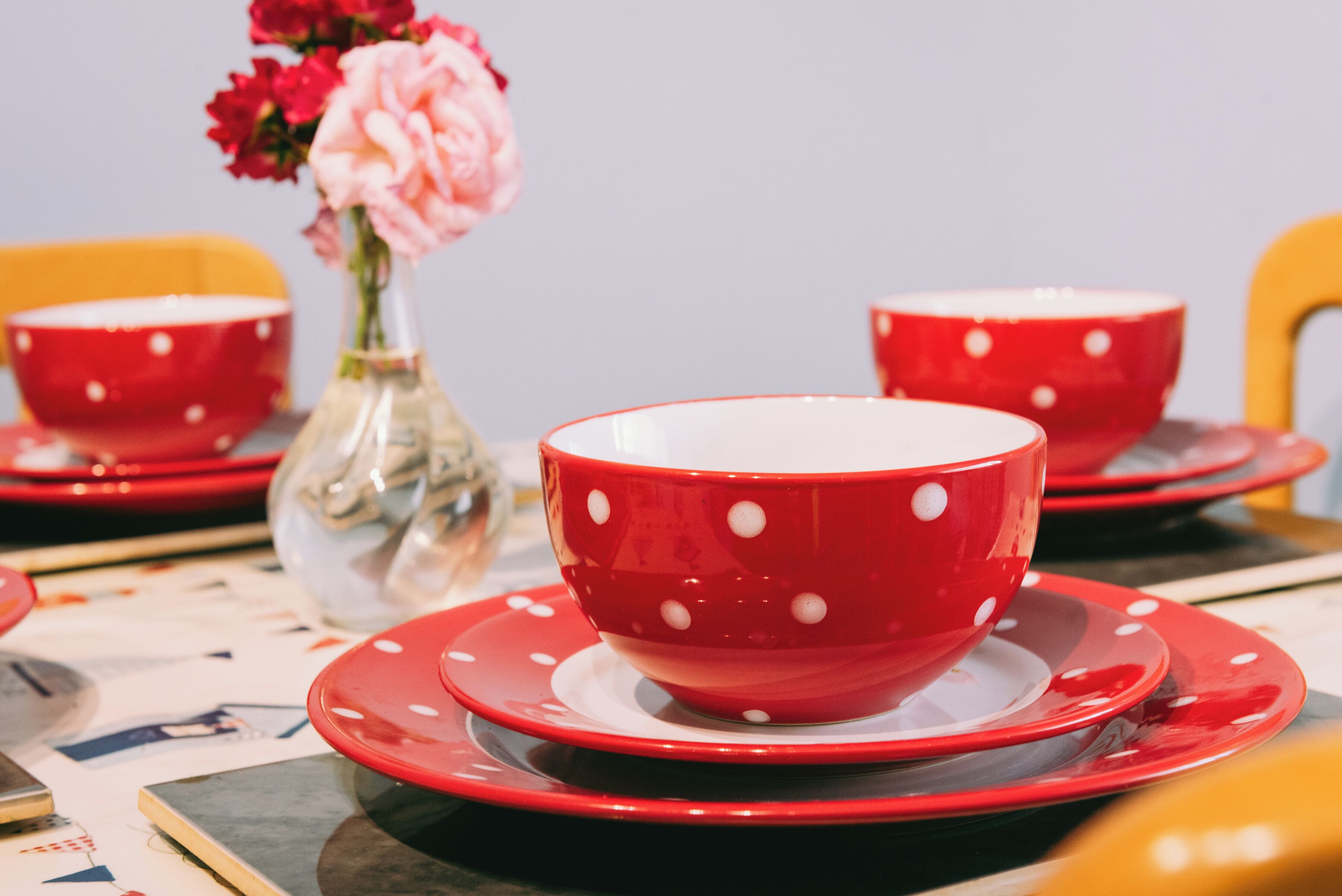 A table with red and white polka-dotted cups and saucers, a clear vase with pink flowers, and a yellow chair in the background.