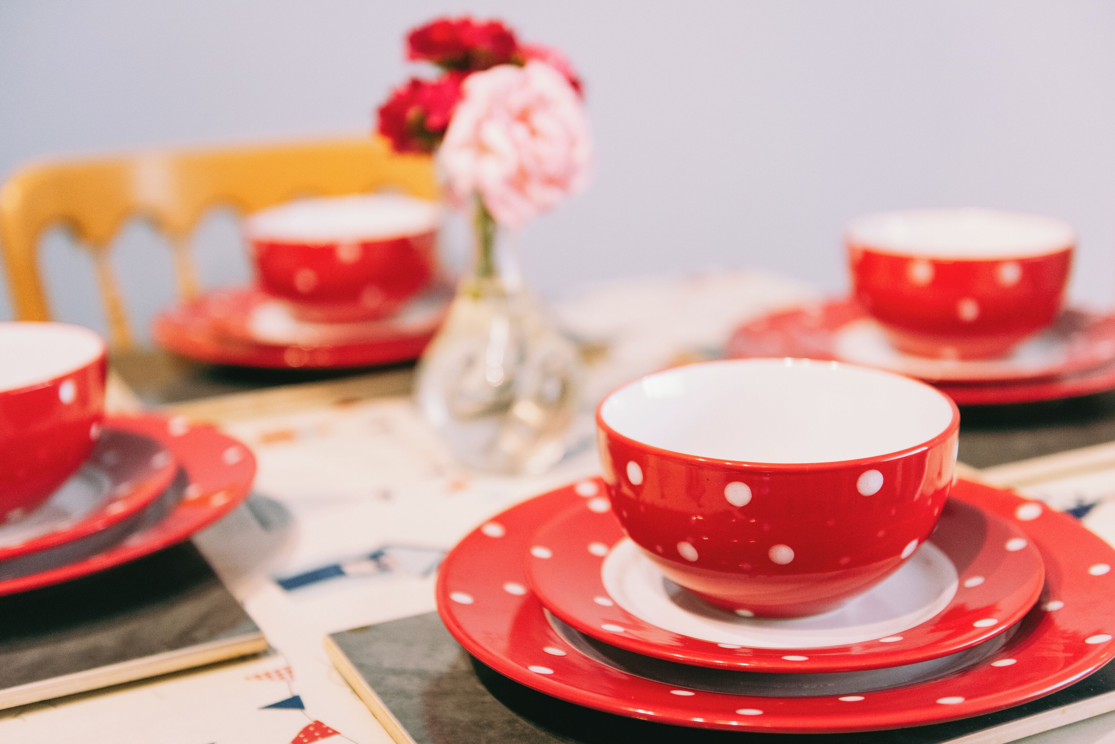Une table dressée avec de la vaisselle rouge et blanche à pois, comprenant des bols et des assiettes, et un vase rempli de fleurs roses.