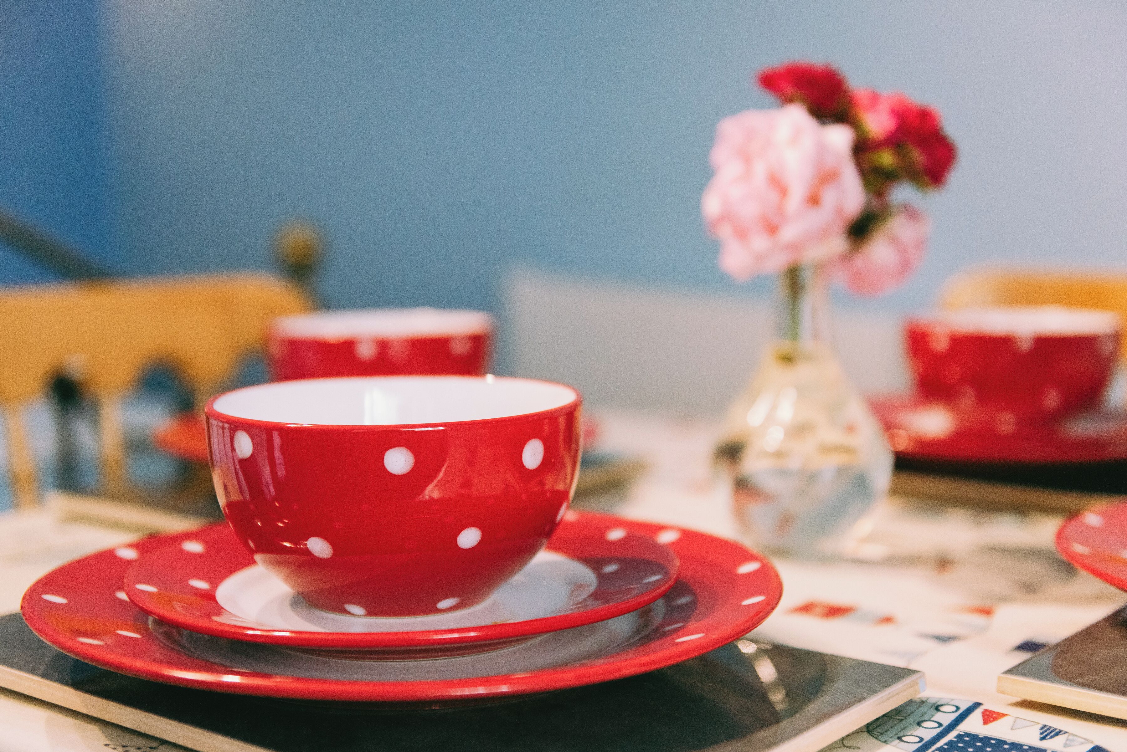 A table with a red polka-dotted cup and saucer, a vase with pink flowers, and a red cup in the background.