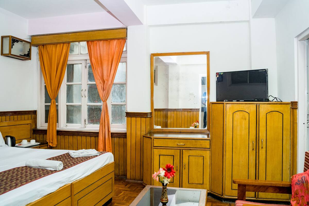 Habitación de hotel con cama, armario de madera, espejo, televisor sobre un mueble bajo y una mesita con un florero.