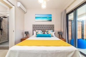 Une chambre dotée d’un grand lit, offrant une vue sur la mer grâce à une porte coulissante, ornée d’un tableau représentant une scène de plage, et équipée d’un système de climatisation moderne.