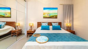 A hotel room with a large bed, two bedside tables, a mirror, and two paintings of a coastal scene. - Immaculate 2-bed Apartment in Pereybere Grand Baie (Grand-Baie)